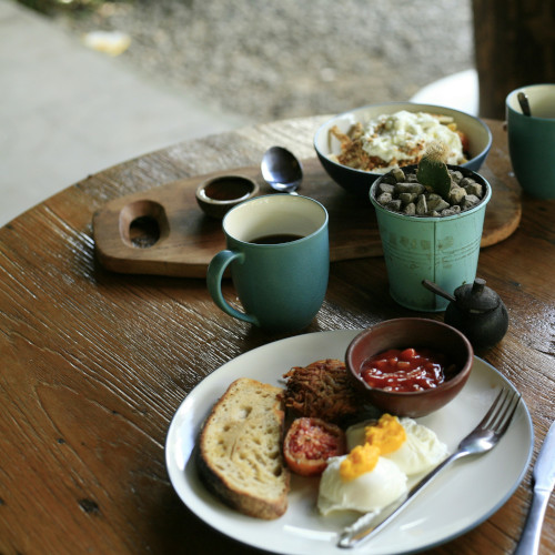 Table de petit-déjeuner avec œufs, pain grillé, saucisses sur assiette, tasses vertes, bols de granola sur table en bois rustique près d'une fenêtre