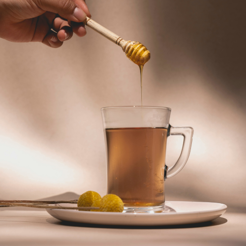 Mano vertiendo miel de cucharón de madera en taza de vidrio con té sobre platillo blanco con rodajas de limón