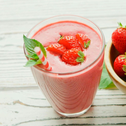 Pink strawberry smoothie in textured glass topped with fresh strawberries and mint leaves on white wooden surface