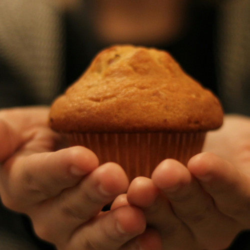Manos sosteniendo muffin recién horneado dorado en capacillo de papel marrón, persona visible borrosa al fondo