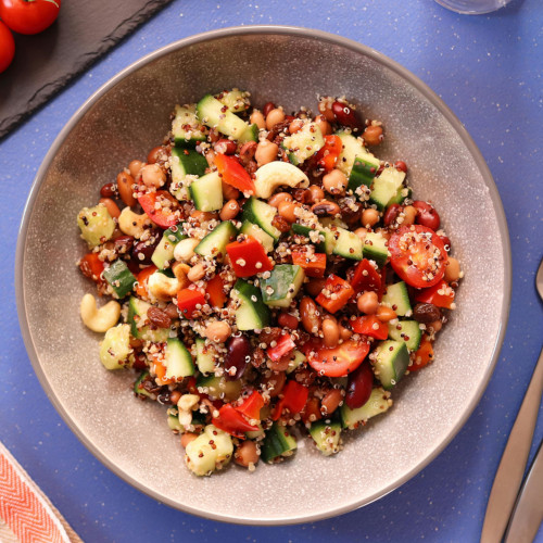 Colorful quinoa salad with chickpeas, tomatoes, cucumber, and cashews in brown speckled bowl on blue surface