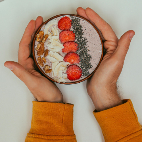 Hands holding smoothie bowl topped with strawberries, banana slices, chia seeds, and walnuts in brown bowl