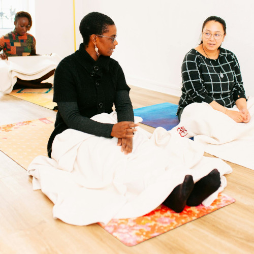 Two women sitting in meditation pose on yoga mats in bright studio, wearing casual clothes during wellness class