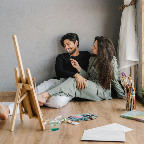 Couple sitting on floor painting together, surrounded by art supplies including easel, brushes, and paint in cozy studio
