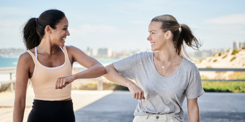 Two smiling women in athletic wear doing elbow bump greeting outdoors with beach and city skyline in background